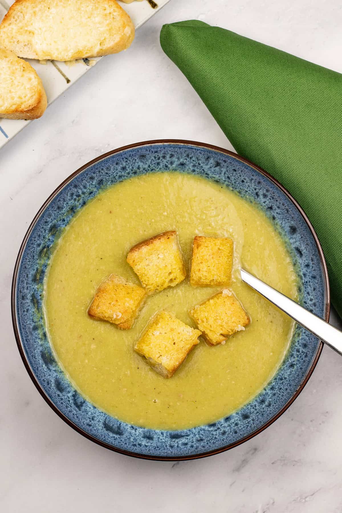Leek and potato soup in a blue patterned bowl with spoon, a plate of cheese on toast and a green napkin on the side.