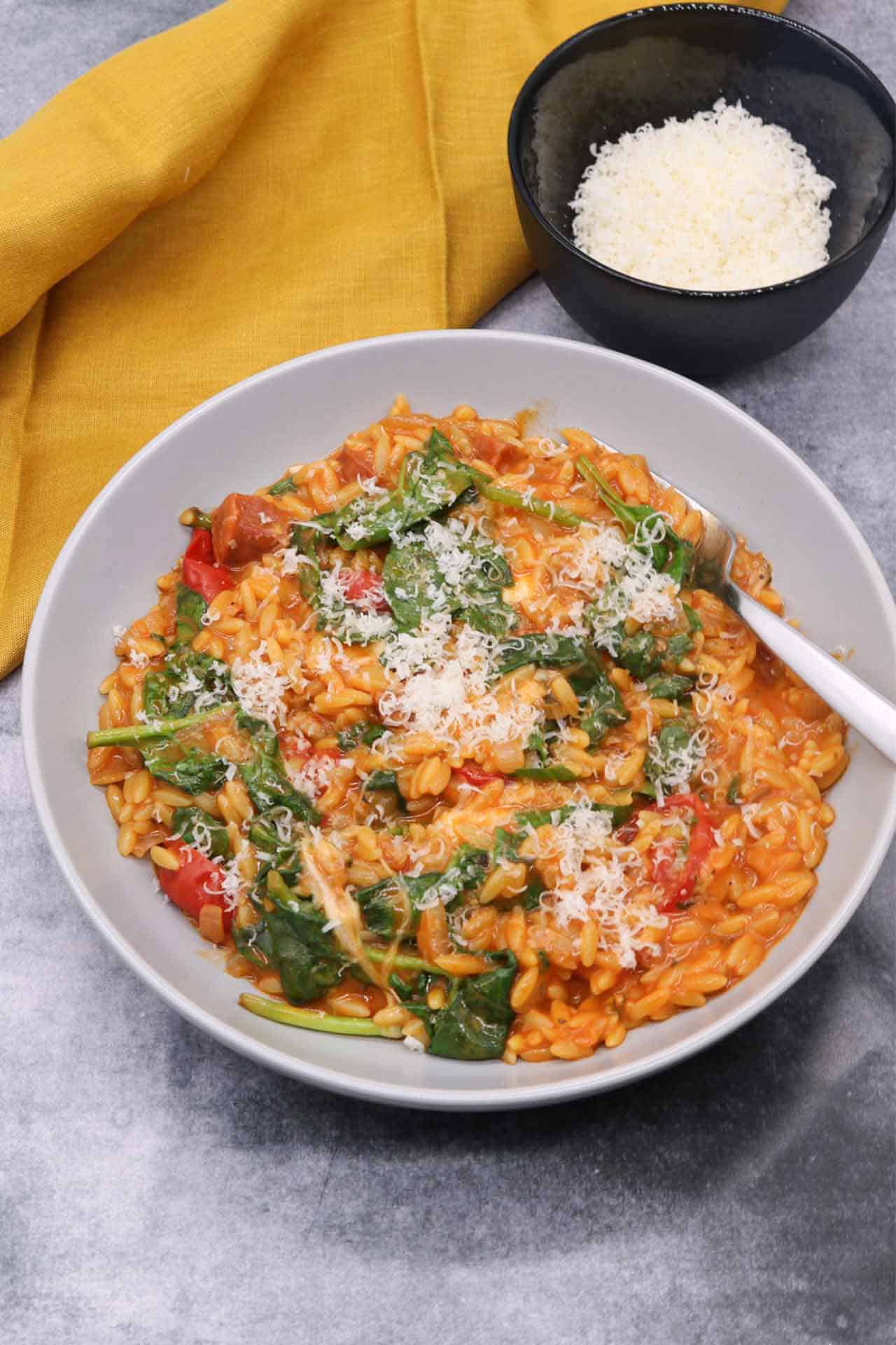 Orzo risotto with chorizo and baby spinach in a grey bowl with fork and small bowl of parmesan and mustard napkin on the side.