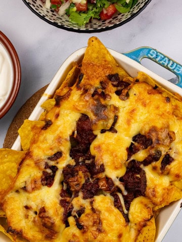 Party nachos with chorizo and spicy cheese sauce in a small rectangular dish with bowls of pico de gallo salsa and sour cream on the side.