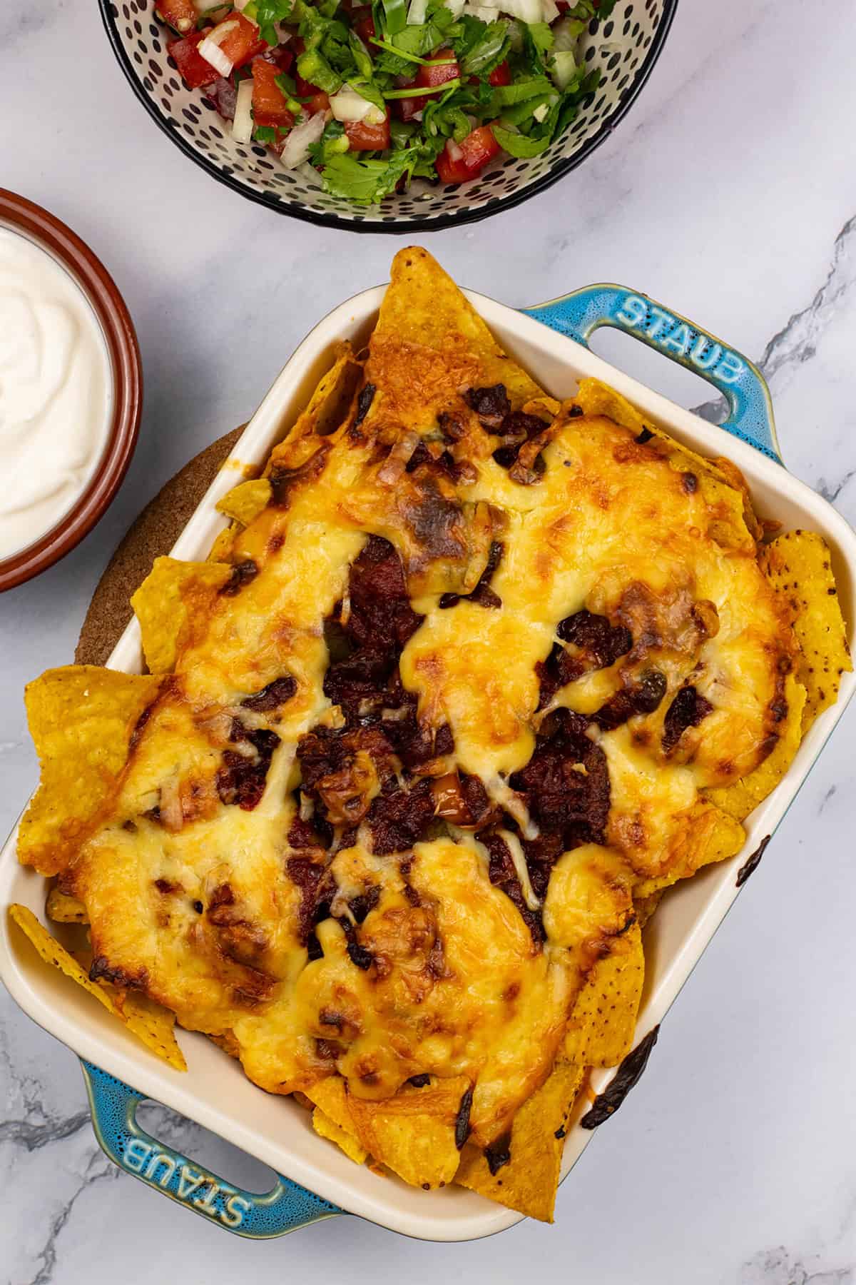 Party nachos with chorizo and spicy cheese sauce in a small rectangular dish with bowls of pico de gallo salsa and sour cream on the side.