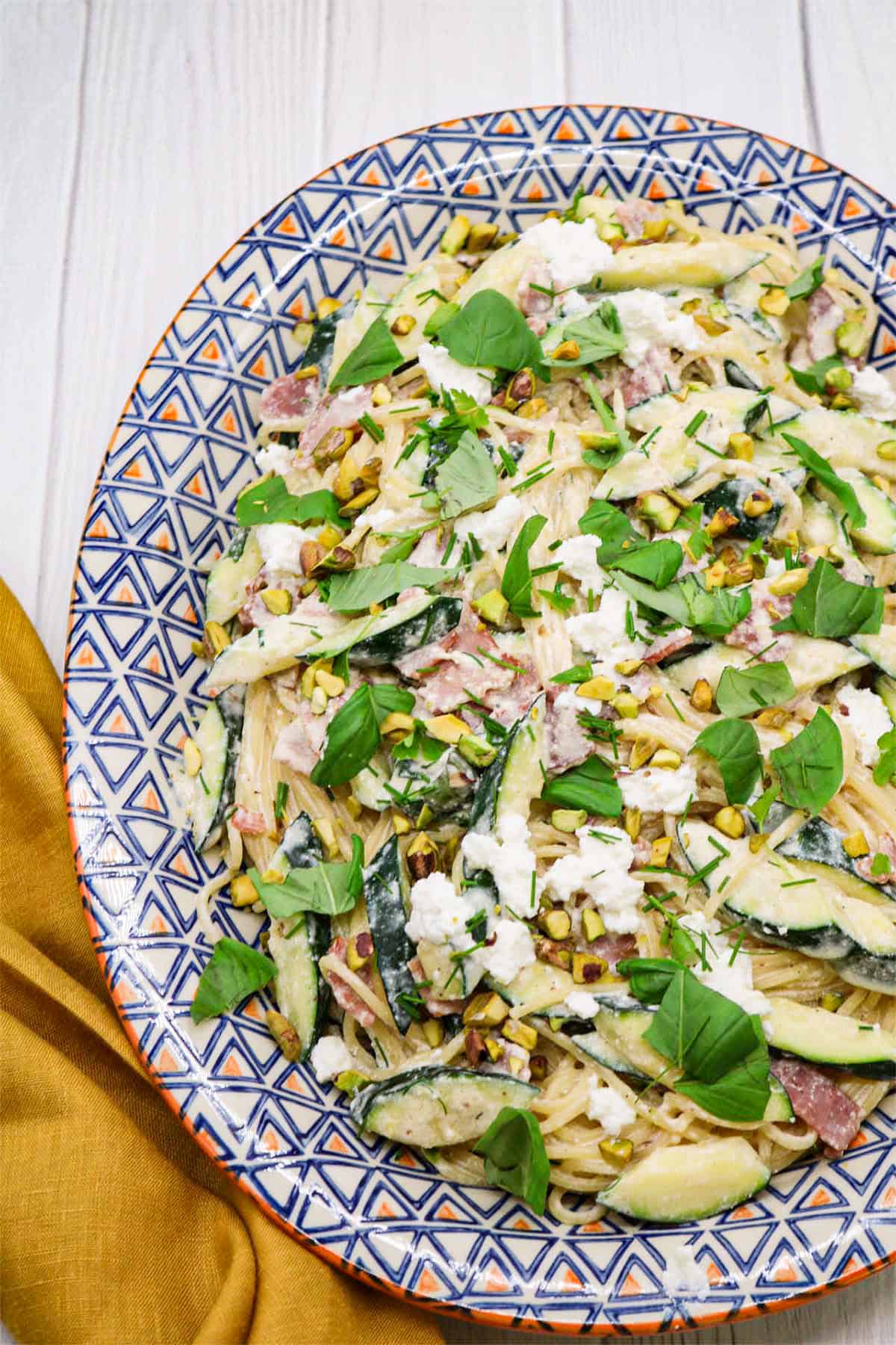 Pasta with crispy bacon, courgette and herbs on an orange, blue and white patterned oval platter.