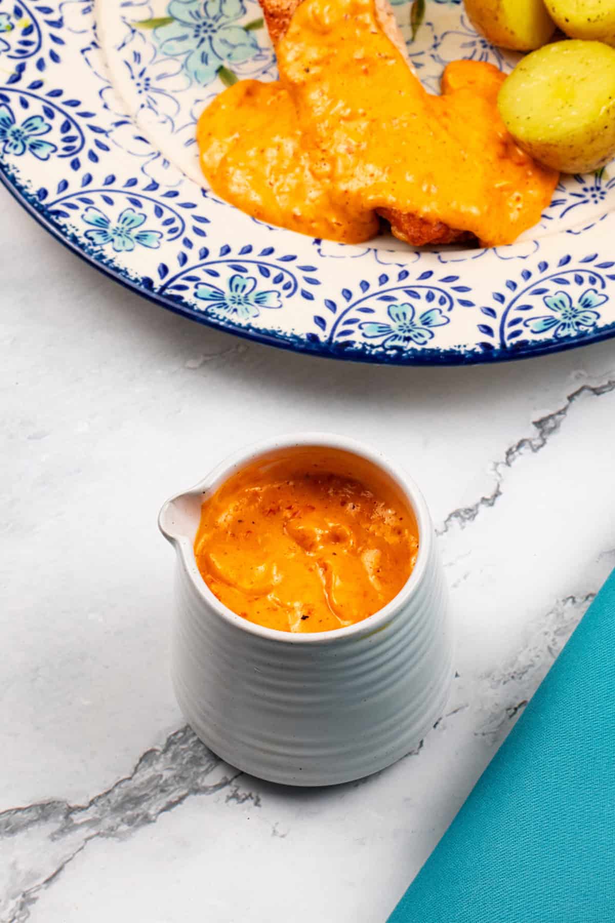 Red pepper and paprika hollandaise sauce made in the Thermomix in a small white jug with a plate of salmon, potatoes and sauce in the background.