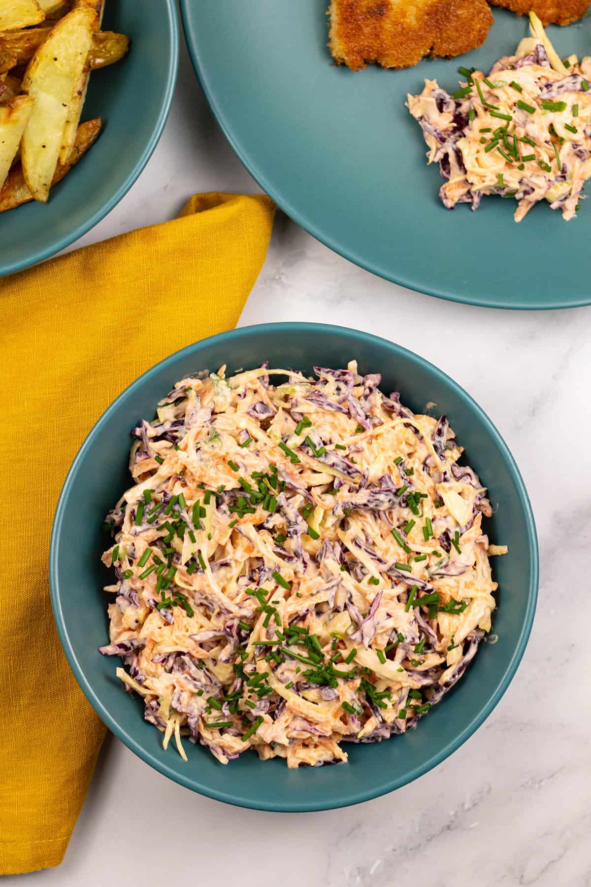 Red and white coleslaw in a teal bowl with a mustard napkin, bowl of chips and dinner plate with pork schnitzel on the side.