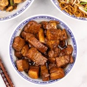 Shanghai-style braised pork belly in an oriental bowl with bowls of chicken and green peppers and fried noodles, and chopsticks on the side.