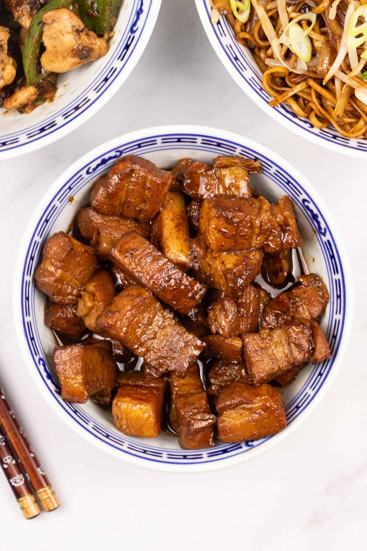 Shanghai-style braised pork belly in an oriental bowl with bowls of chicken and green peppers and fried noodles, and chopsticks on the side.