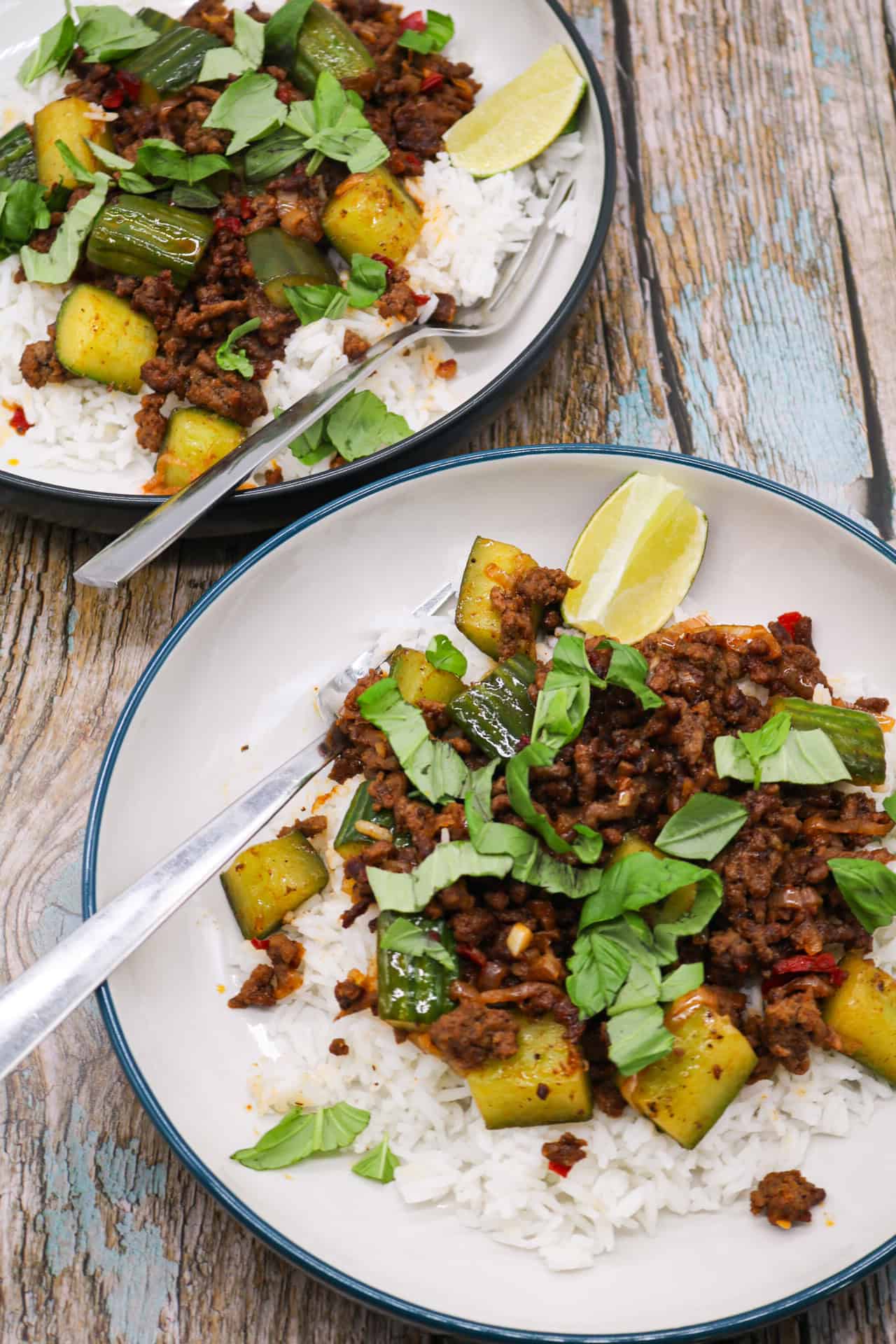 Two bowls of spicy beef and crushed cucumber stir-fry served on rice with forks and lime wedges.