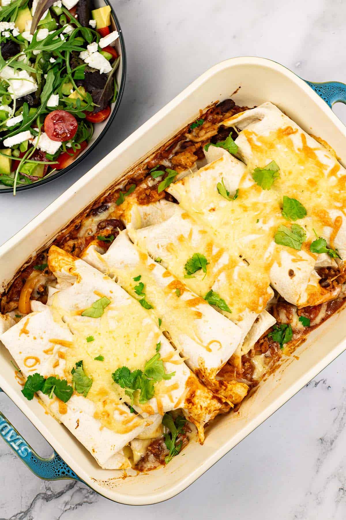 Turkey enchiladas in a large rectangle oven dish with a bowl of salad on the side.