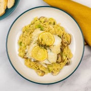 Baked cod with creamy mushroom and leek orzo in a bowl with a plate of sliced baguette and a mustard napkin on the side.