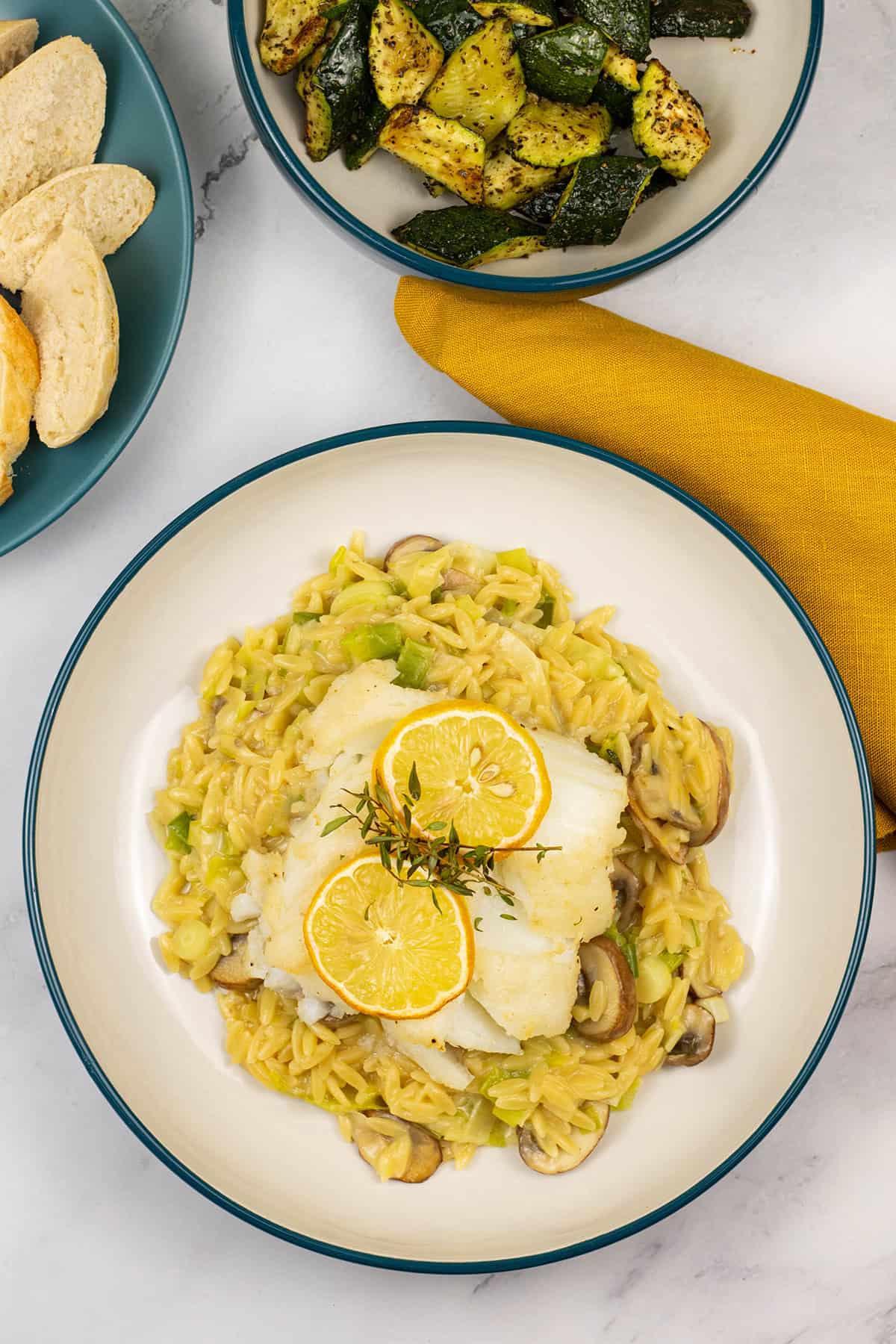 Baked cod with creamy mushroom and leek orzo in a bowl with a plate of sliced baguette and a bowl of air fryer courgettes on the side.