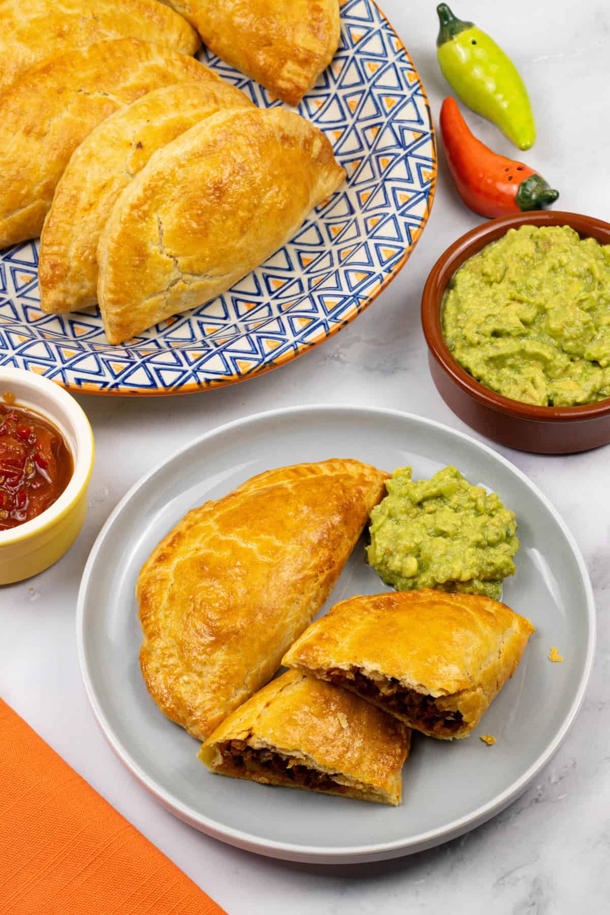 2 beef and chorizo empanadas on a grey side plate with some guacamole, a platter of empanadas and dishes of guacamole and chilli and tomato jam on the side.