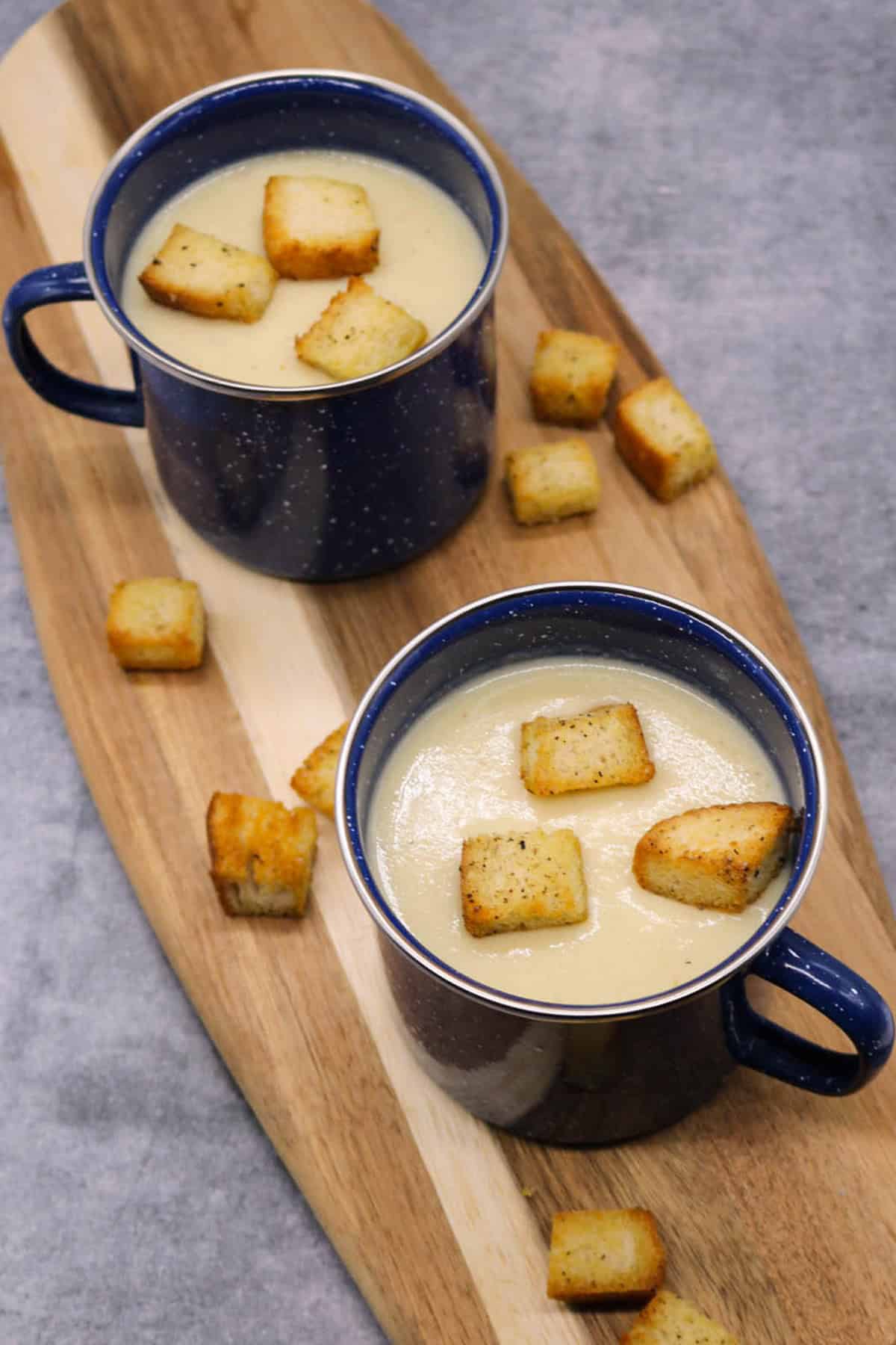 Cauliflower cheese soup in 2 blue mugs sitting on a board with croutons on the side.