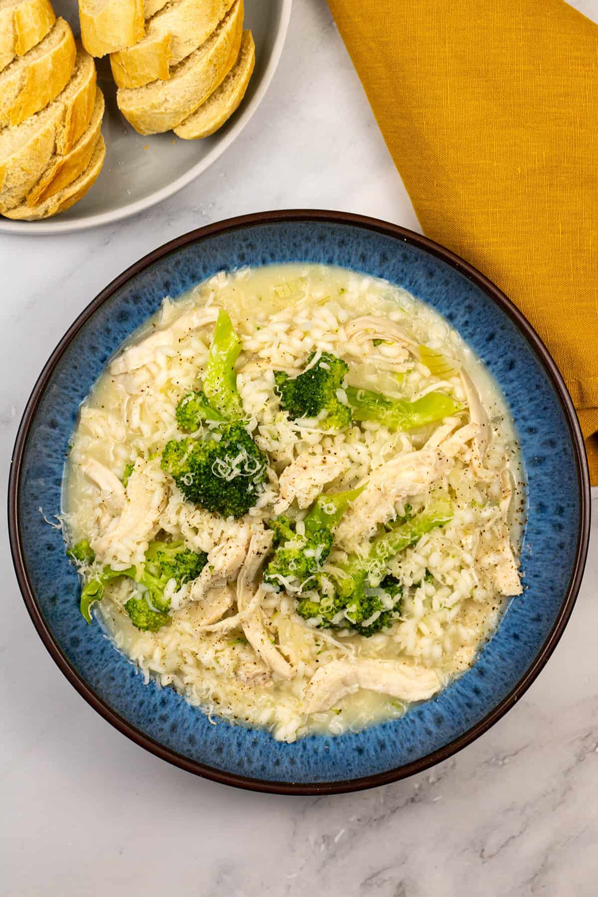 Chicken and broccoli risotto in a blue patterned bowl with a plate of sliced baguette and a mustard napkin on the side.