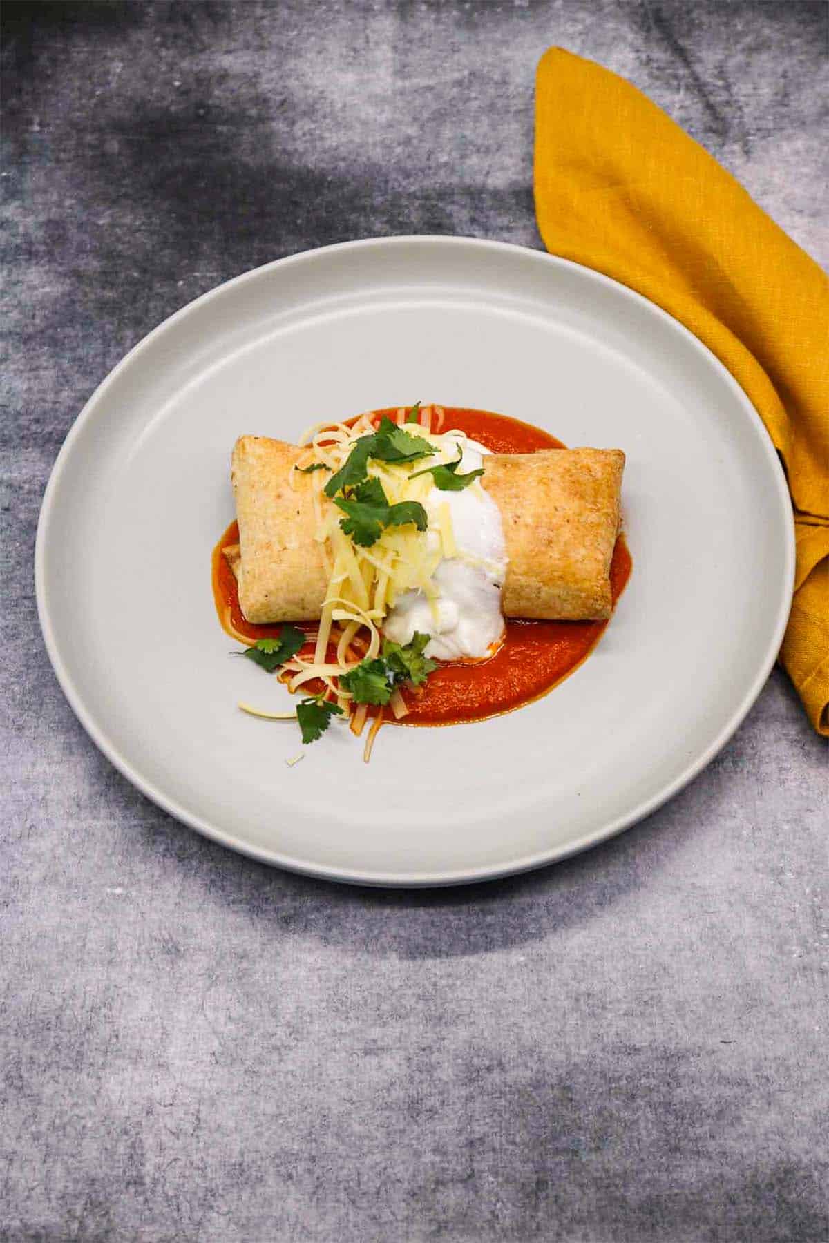 Chicken chimichanga on grey plate covered in cheese, coriander and soured cream on top of tomato sauce
