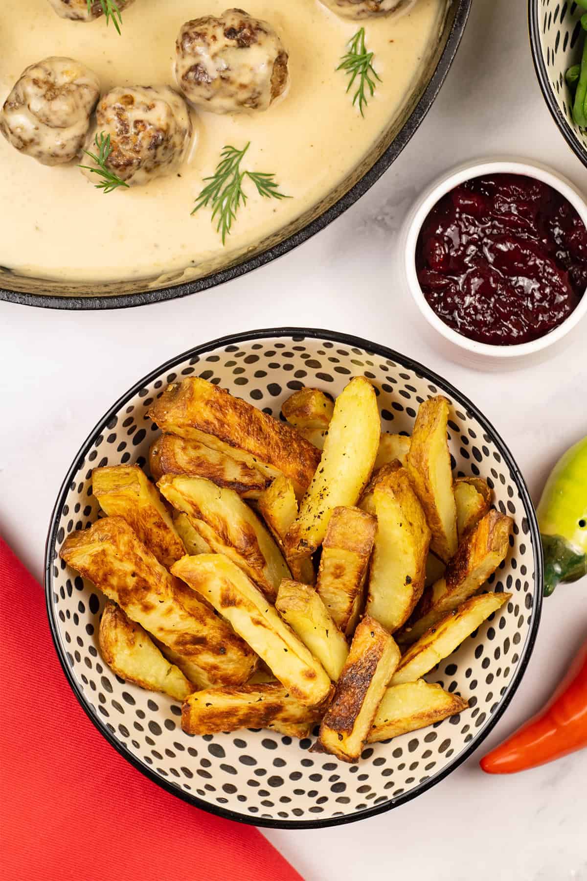 Chunky oven chips in a small serving bowl with scandi-style meatballs with gravy and a small dish of lingonberry spread on the side.