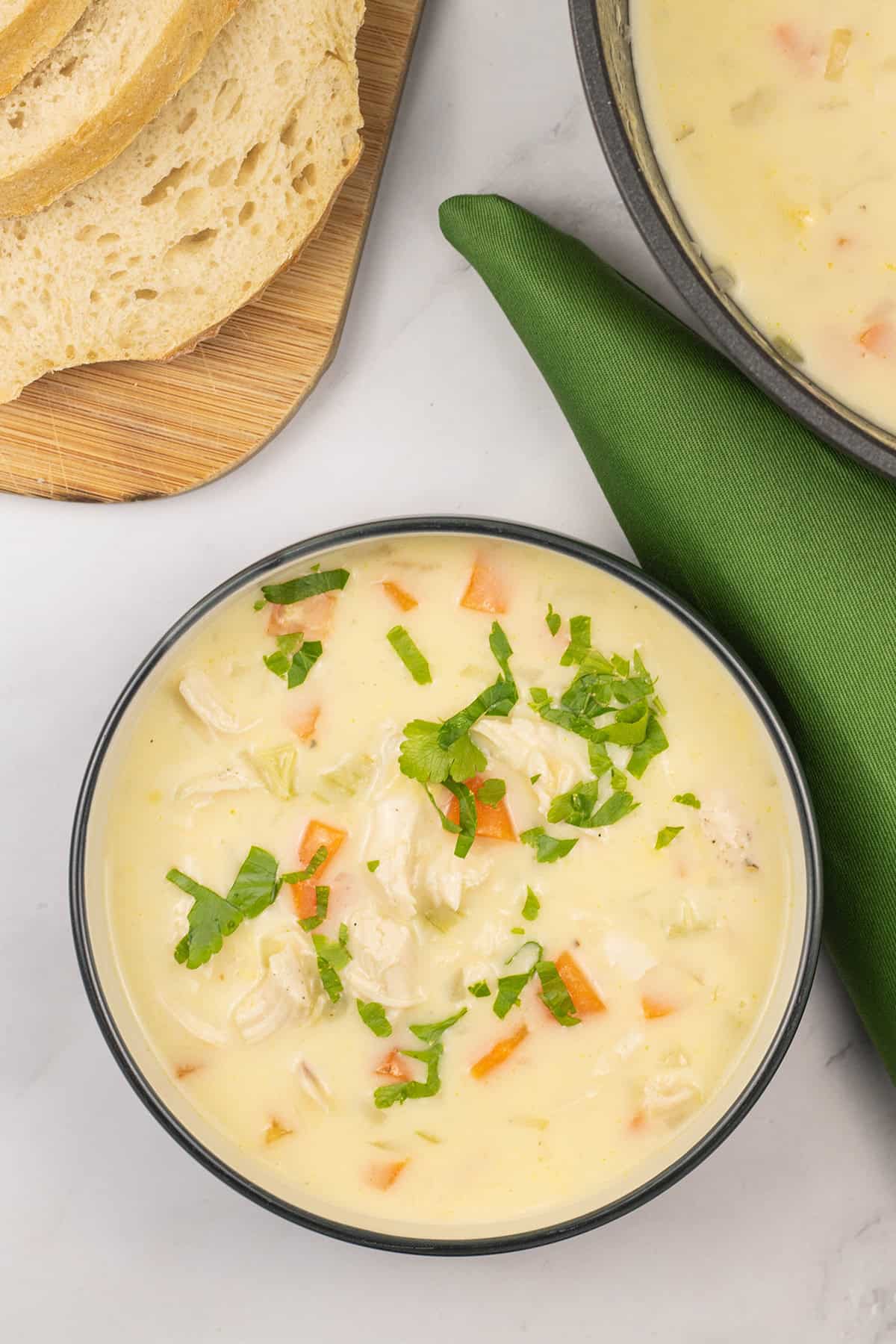 Cream of chicken and vegetable soup in bowl with a board of sliced bread, soup pot and green napkin on the side.