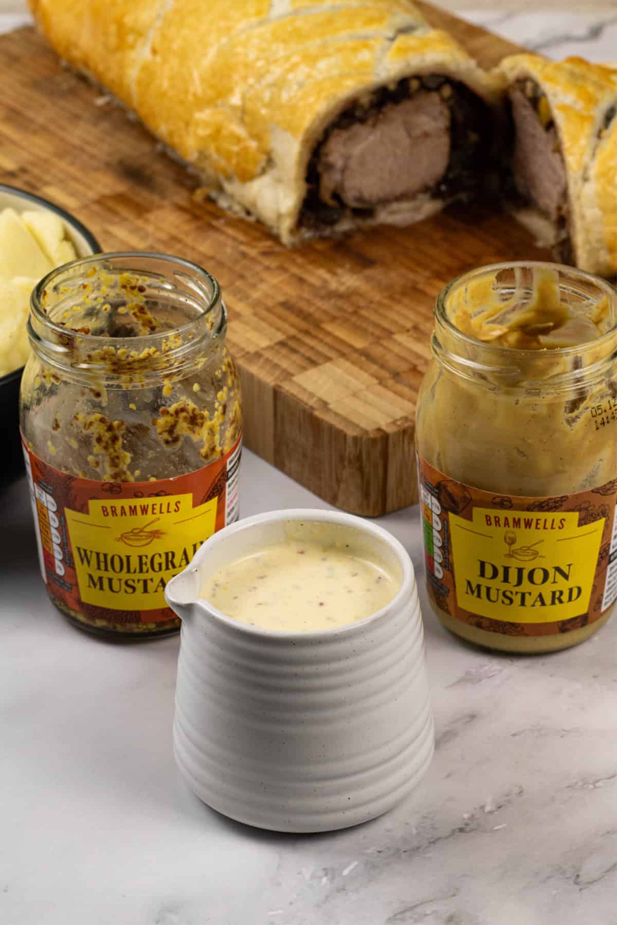 A small jug of easy mustard cream sauce with jars of wholegrain and dijon mustard on the side and a pork wellington with black pudding in the background.