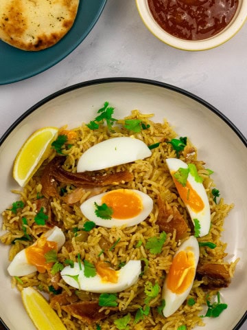 A portion of kedgeree in a black and cream bowl with a plate of naan and small dish of mango chutney on the side.