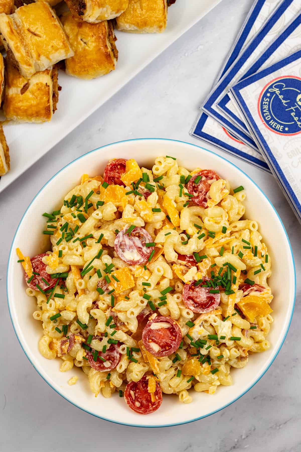 Mac 'n' cheese salad in a round serving bowl with a platter of sausage rolls and paper napkins on the side.