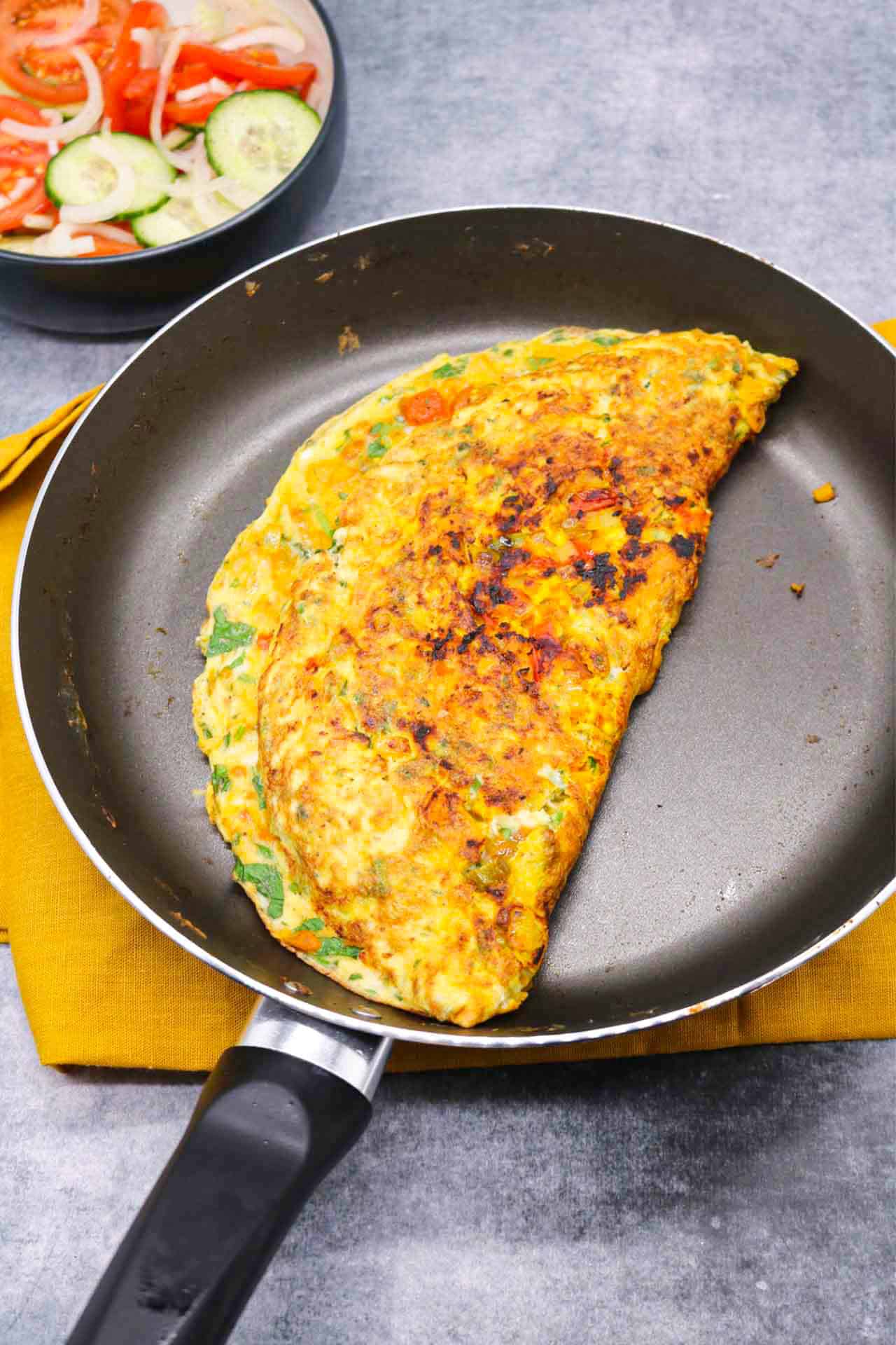 Masala omelette in frying pan with a bowl of tomato and cucumber salad on the side