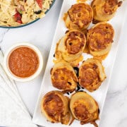 Pepperoni pizza rolls on a rectangle platter with ramekin of marinara sauce, bowl of pasta salad and paper napkins on the side.
