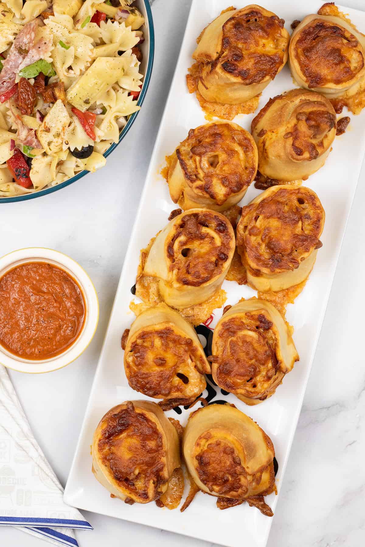 Pepperoni pizza rolls on a rectangle platter with ramekin of marinara sauce, bowl of pasta salad and paper napkins on the side.
