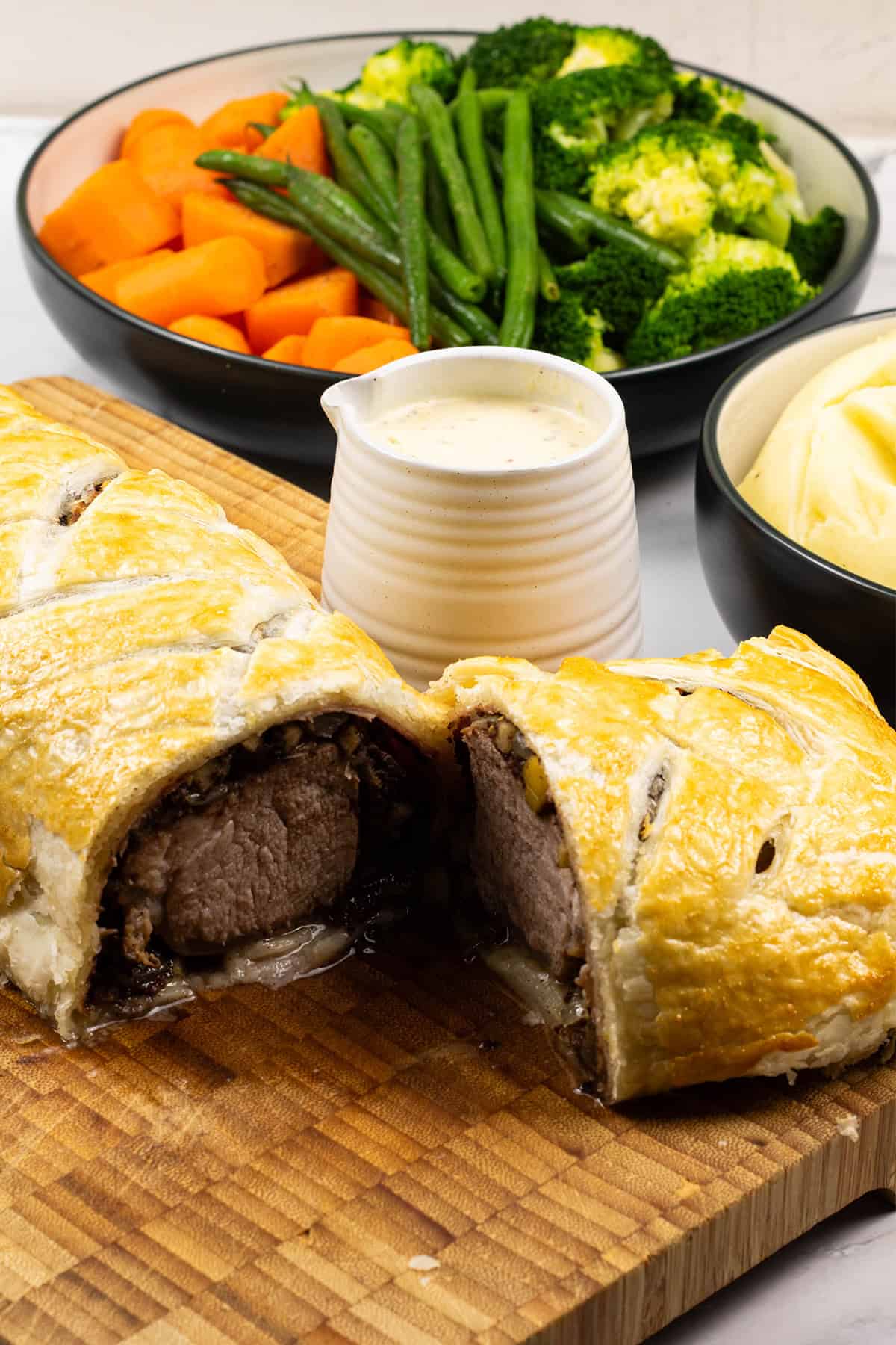 Pork wellington with black pudding cut open on wooden board with a small jug of mustard cream sauce and bowls of mash and vegetables on the side.