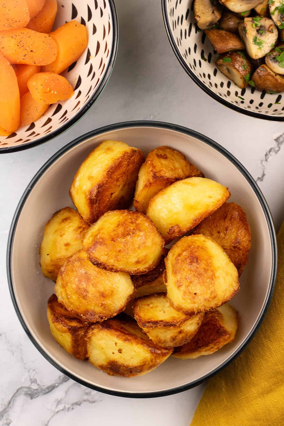 Roast potatoes in a black and cream serving bowl with bowls of carrots and garlic mushrooms on the side.