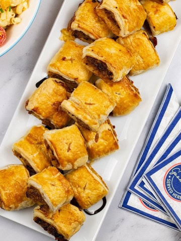 Sausage rolls on a rectangular platter with a bowl of mac 'n' cheese salad and paper napkins on the side.