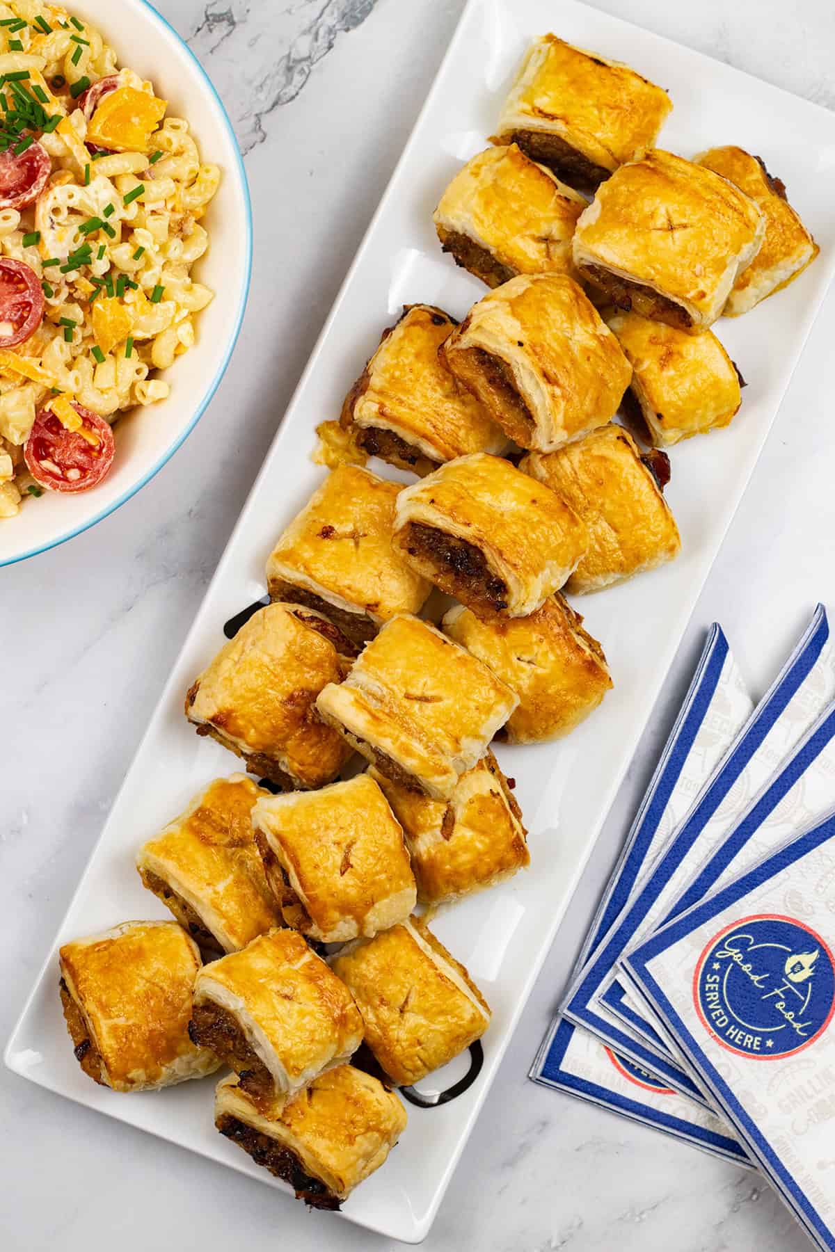 Sausage rolls on a rectangular platter with a bowl of mac 'n' cheese salad and paper napkins on the side.