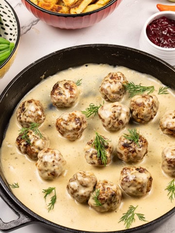 Scandi-style meatballs with gravy in a skillet, with dishes of chips, green beans and lingonberry spread on the side.