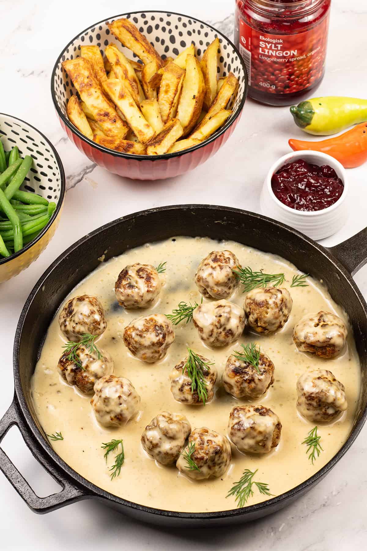 Scandi-style meatballs with gravy in a skillet, with dishes of chips, green beans and lingonberry spread on the side.