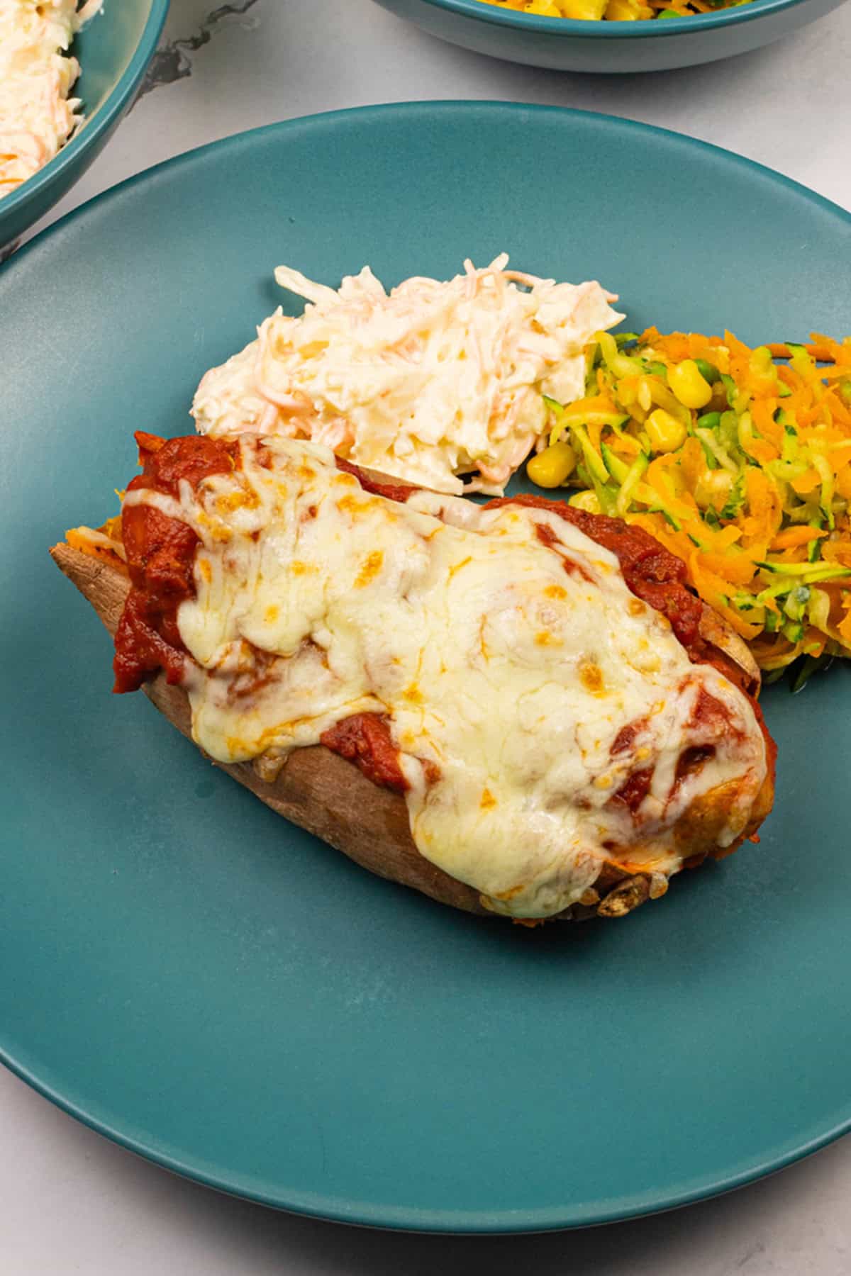 Close up of sweet potato with sausage meatball marinara, coleslaw and carrot salad on a teal dinner plate.