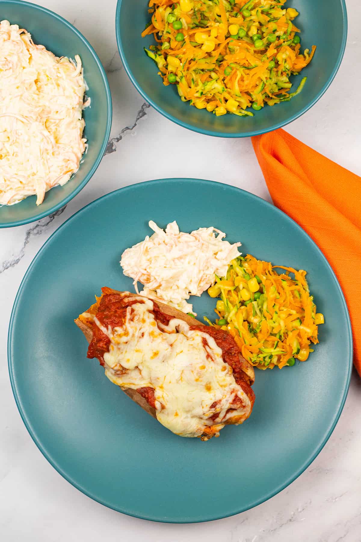 Sweet potato with sausage meatball marinara, coleslaw and carrot salad on a teal dinner plate with bowls of coleslaw and carrot salad on the side.