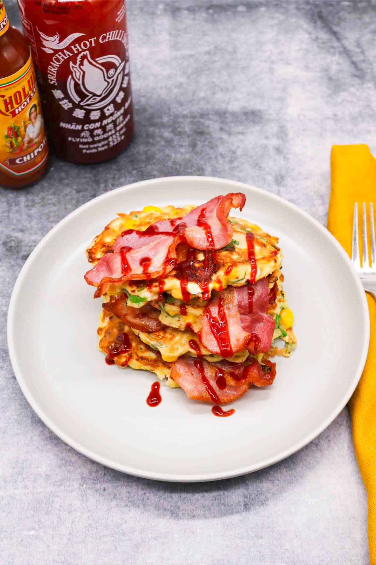 Sweetcorn Feta and Spring onion pancakes stacked on a grey plate with bacon and drizzle of Sriracha