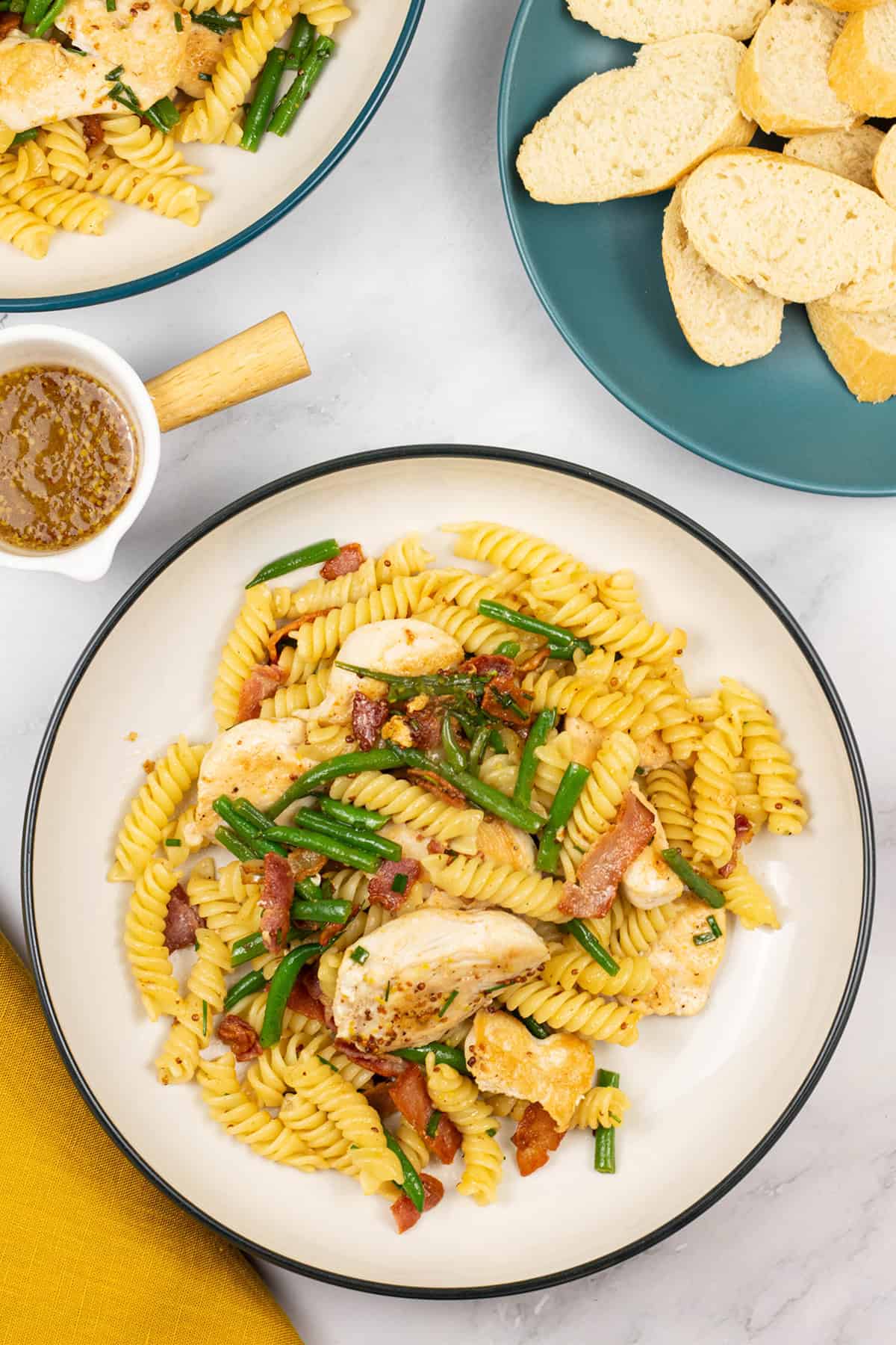 Two bowls of warm chicken and bacon pasta salad with a small jug of dressing and plate of sliced baguette on the side.