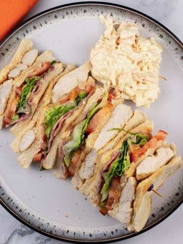 A club sandwich and coleslaw on a dinner plate with an orange napkin on the side.