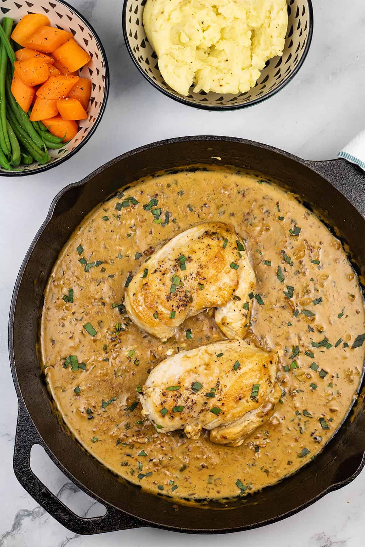 Creamy tarragon chicken in a skillet with bowls of mashed potatoes, and carrots and green beans on the side.