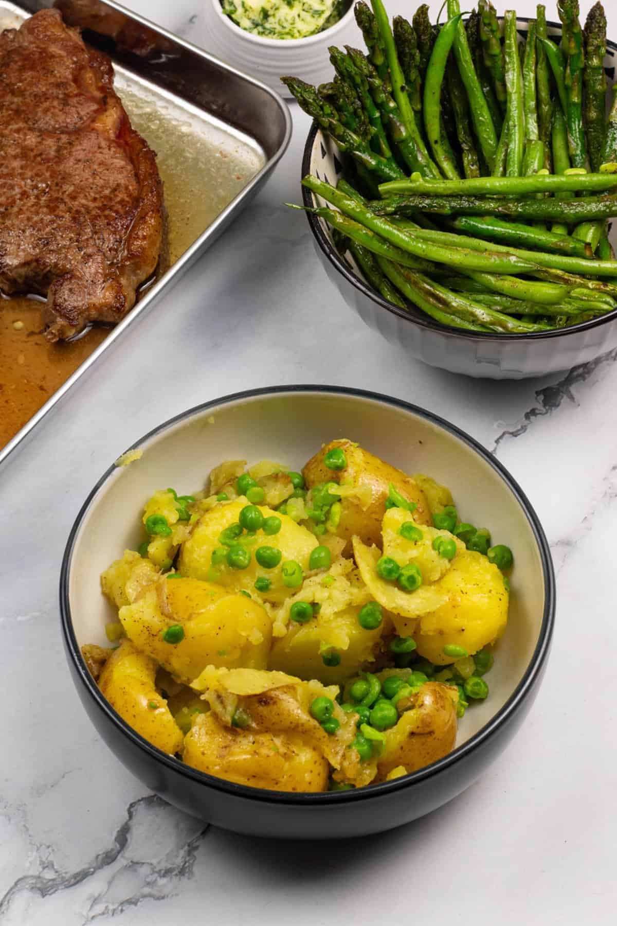 Easy crushed potatoes with peas in a black and cream serving bowl with a bowl of sautéed asparagus and green beans, and a tray with sirloin steaks on the side.