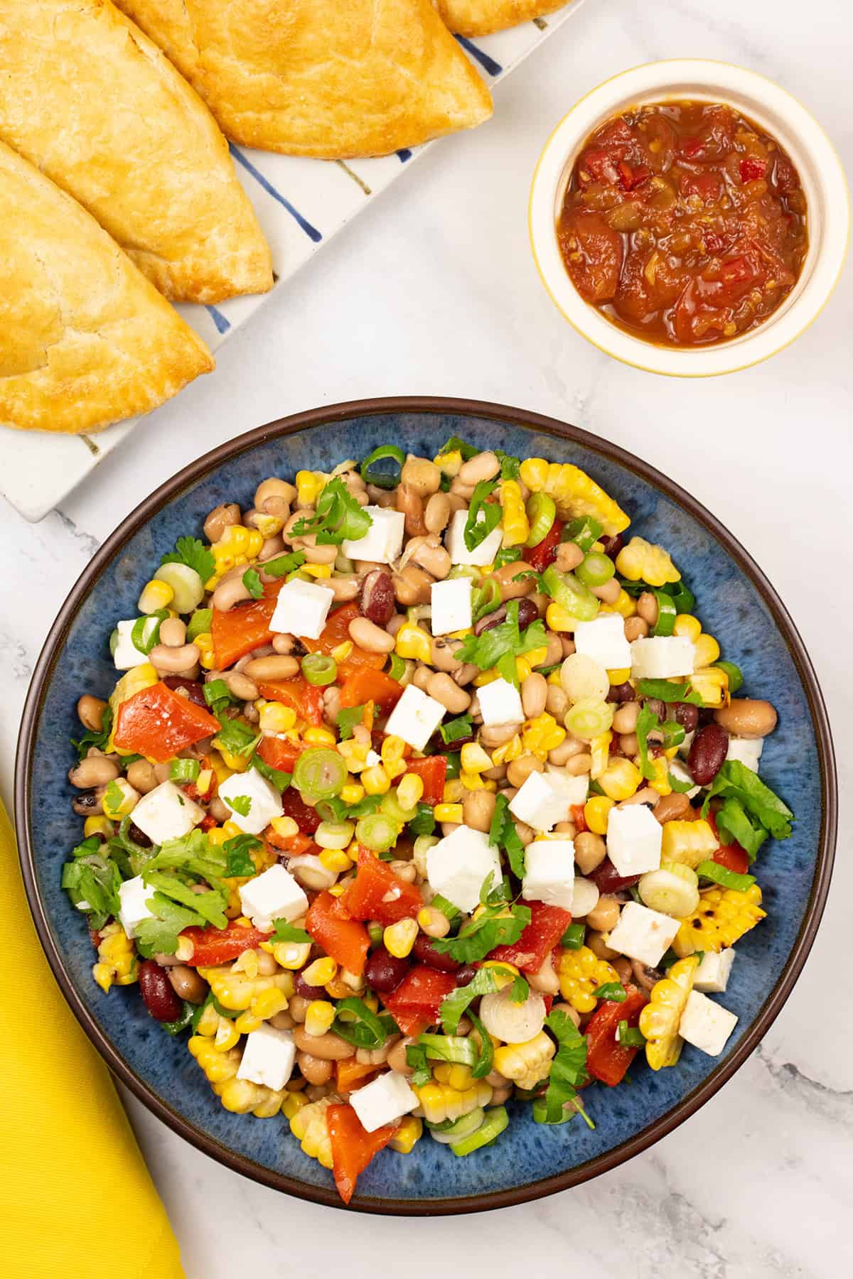 Grilled sweetcorn salad with feta in a blue serving bowl with a platter of empanadas and small dish of chilli and tomato jam on the side.