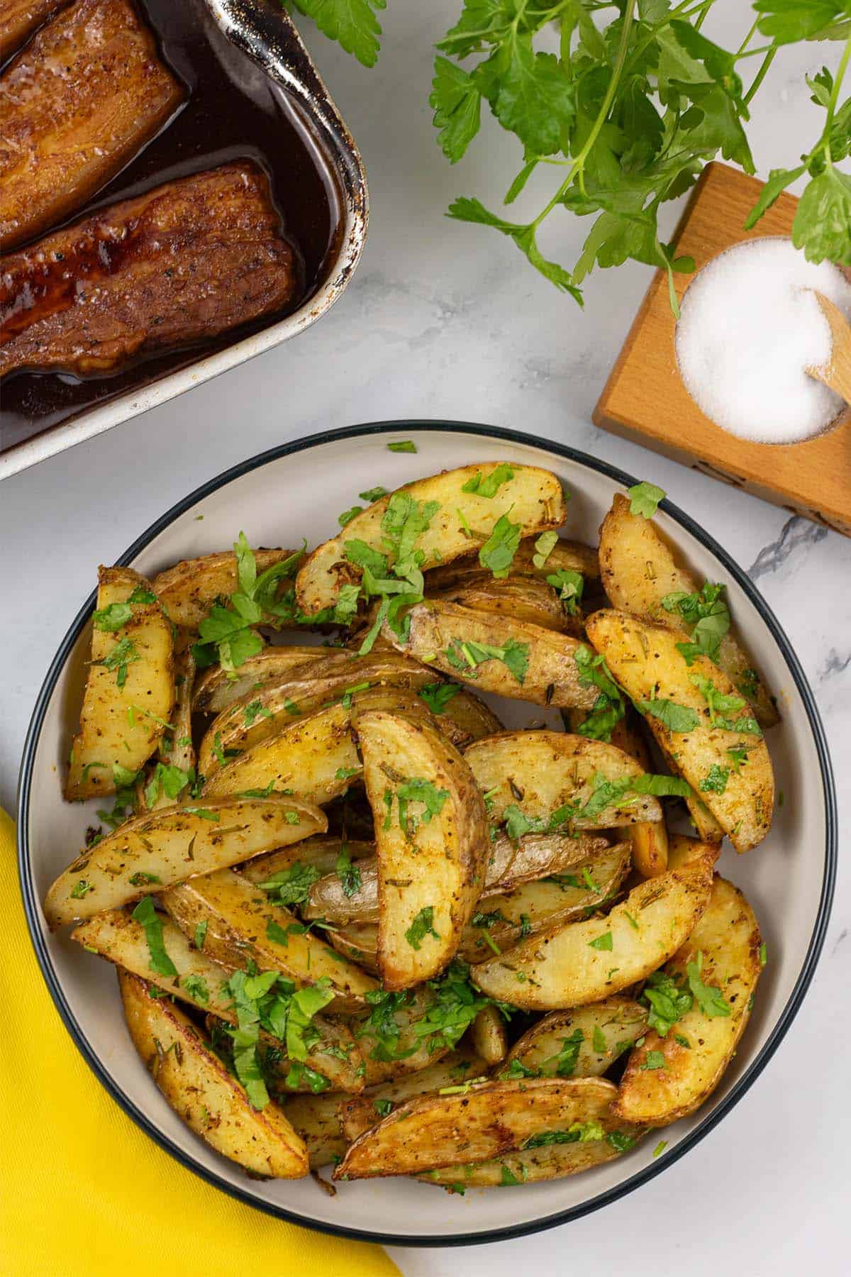 Herby garlic butter potato wedges in a black and cream serving bowl with a tin of pork belly slices and yellow napkin on the side.