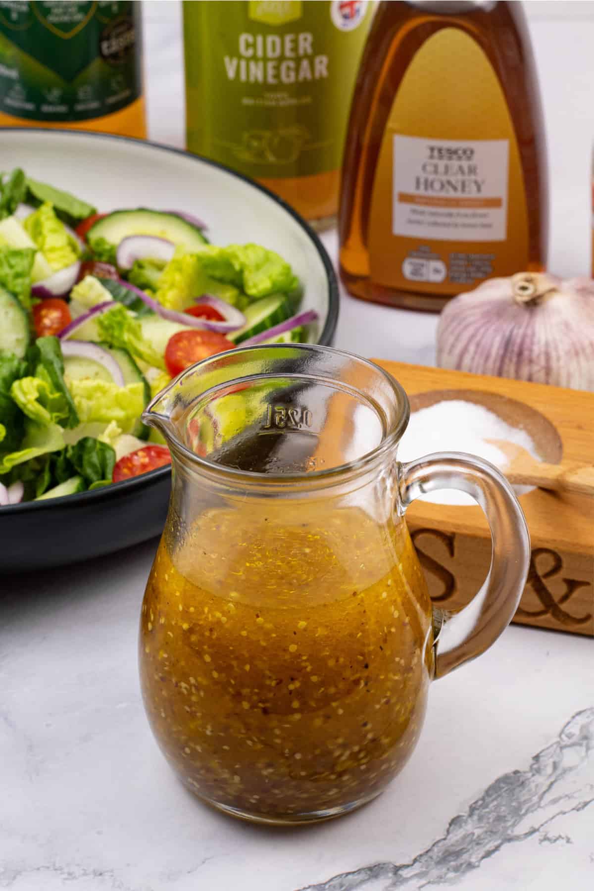 Honey mustard dressing in a small glass jug with a bowl of salad and the ingredients in the background.