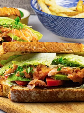 2 hot smoked salmon club sandwiches on a board with a bowl of chunky oven chips in the background.