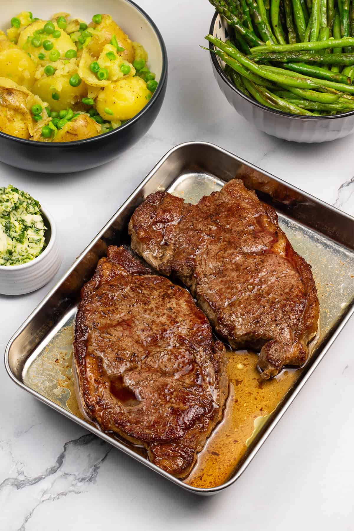 Perfect sirloin steaks resting on a tray, with bowls of crushed potatoes and peas, sautéed asparagus and green beans, and a small dish of wild garlic and rosemary butter on the side.