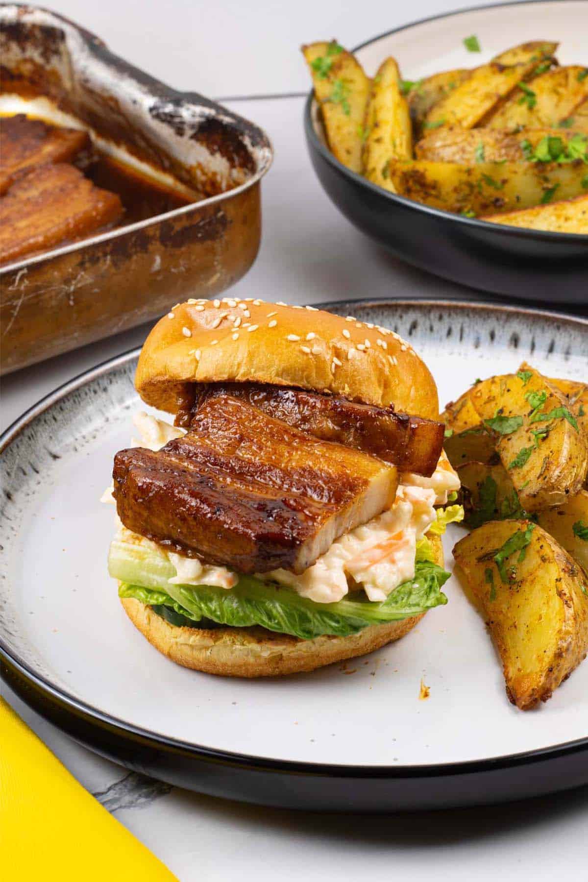 Pork belly slices in BBQ sauce in a brioche bun with salad leaves and coleslaw, with herby garlic butter potato wedges.