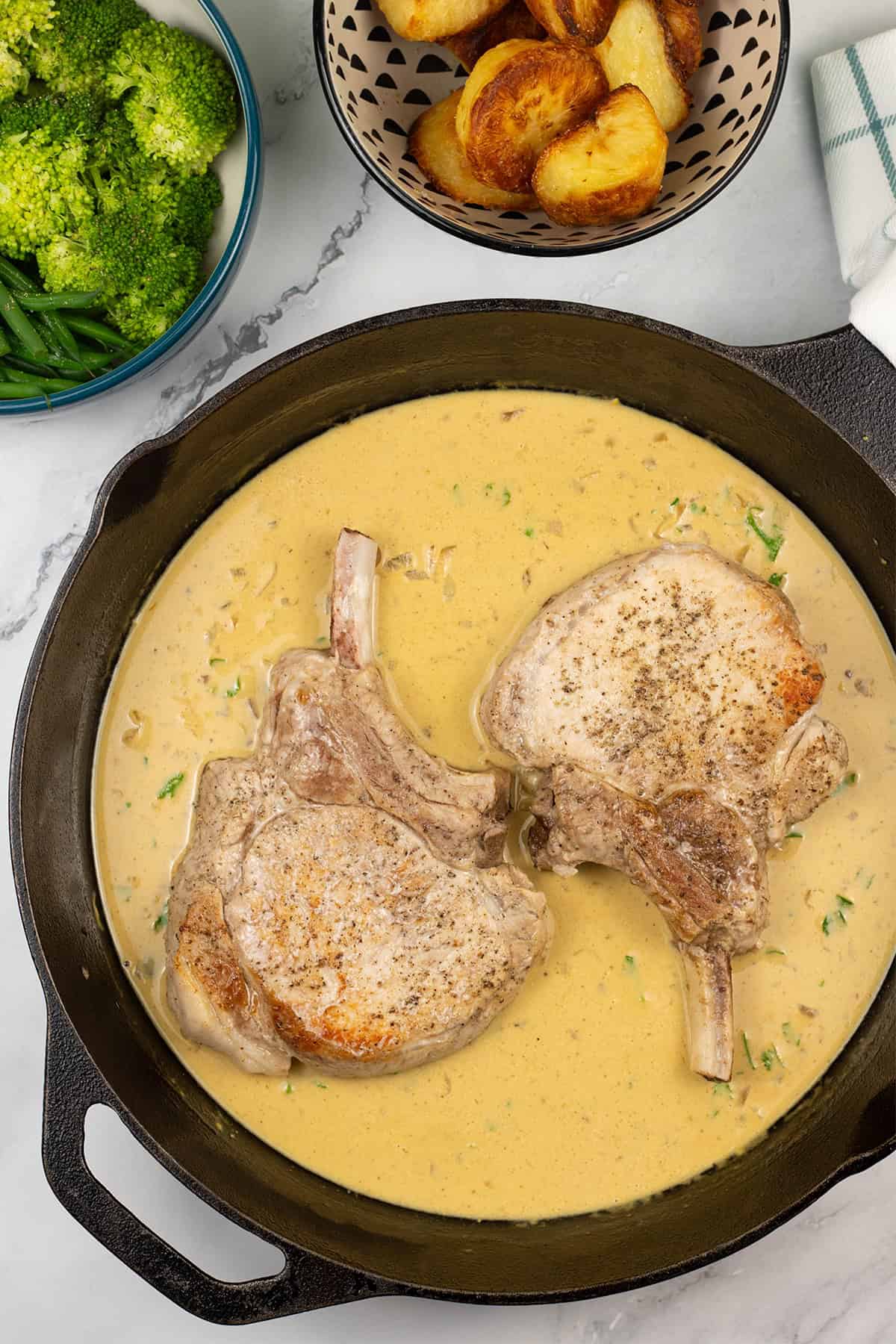 2 pork chops with dijon sauce in a skillet pan with bowls of roast potatoes, green beans and broccoli on the side.