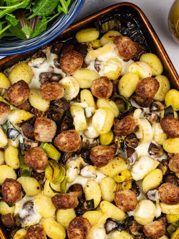 Sausage and taleggio gnocchi bake on a large baking tray with a bowl of baby salad leaves and a small jug of Italian dressing on the side.