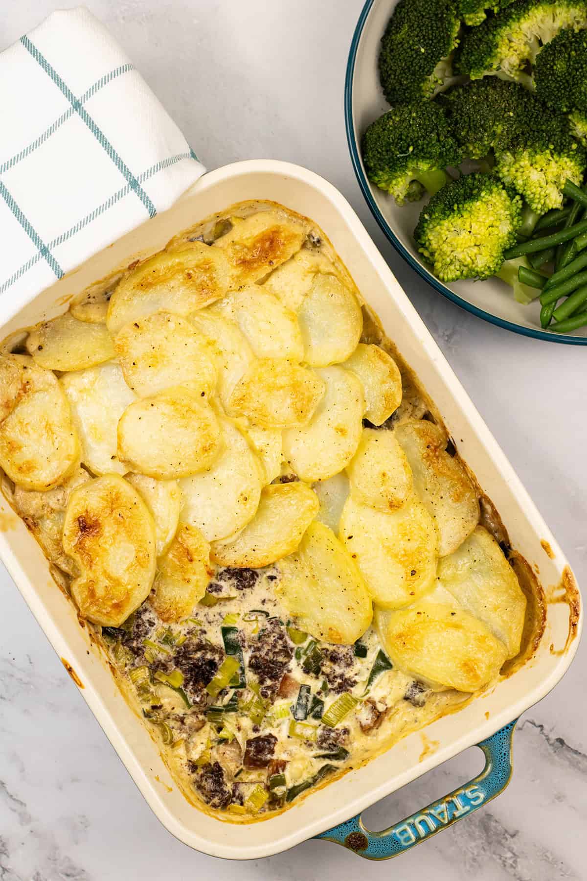 Smoked haddock, black pudding and leek gratin in a rectangle Staub dish with a bowl of broccoli and green beans on the side.