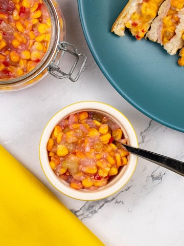 Sweetcorn relish with chilli in a yellow ramekin plus a large jar in the background with a plate with a toastie.