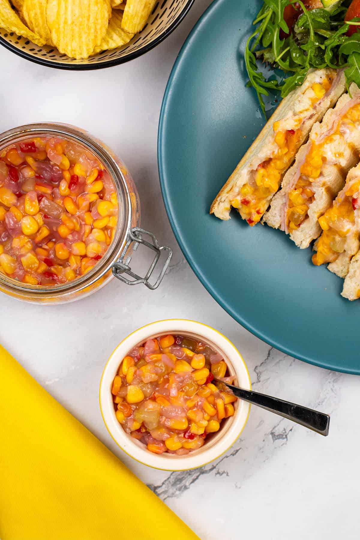 Sweetcorn relish with chilli in a yellow ramekin plus a large jar in the background with a bowl of crisps and a plate with a toastie and salad.