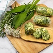 3 logs of wild garlic and rosemary butter on a board with sprigs of rosemary and leaves and flowers of wild garlic on the side.